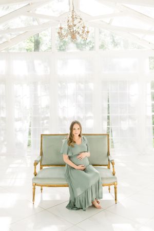 A maternity portrait in an elegant white sunroom with a chandelier and vintage settee.