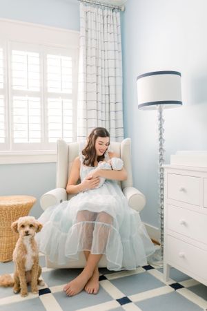 Cozy nursery corner with white armchair, floor lamp, and small dog looking up at the camera.