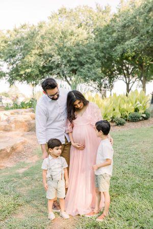 A family gathers outdoors in casual wear with a pregnant mother in a pink dress.