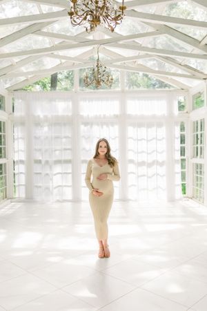 Elegant maternity portrait in a light-filled conservatory featuring a fitted cream dress.