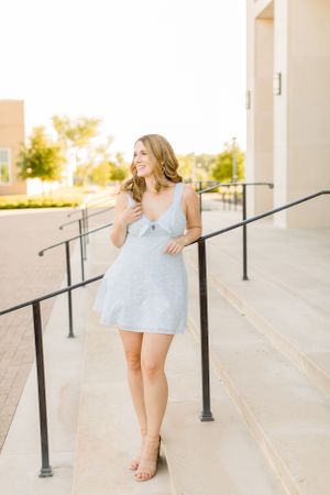 Woman wearing blue dress stands outdoors against architectural backdrop with handrails.