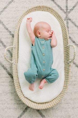 Sleeping baby in teal button-up romper stretching in woven bassinet.