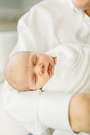 Peaceful sleeping baby wrapped in white swaddle against light background.