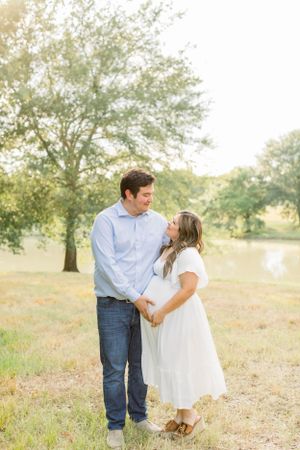 A romantic outdoor portrait in a sunlit field with trees and a pond in the background.