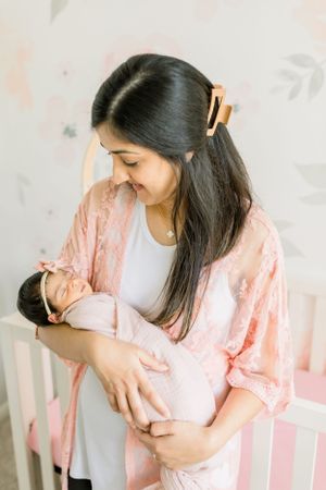 New parent in pink robe tenderly holds and gazes at sleeping newborn in nursery with floral wallpaper.