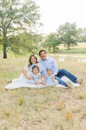 Family portrait on a white blanket in a sunny field with trees and water in the background.