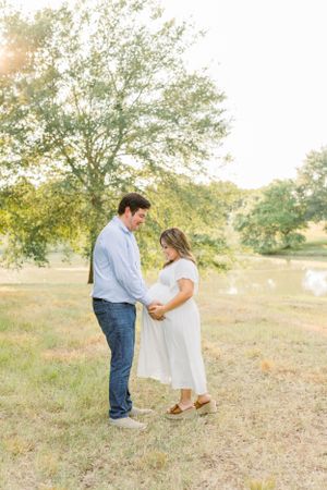 Couple sharing intimate moment in field with sunlit trees and pond in background.