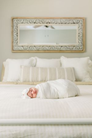Swaddled baby lying on white bedding beneath ornate mirror in serene bedroom setting.