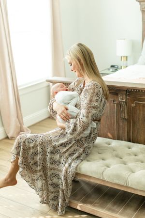 A tender moment as a parent in a floral dress cradles a newborn baby on a tufted bench beside a window.