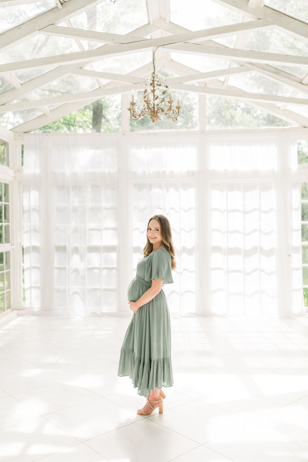 Expectant mother in sage green dress poses in an airy white sunroom with crystal chandelier.