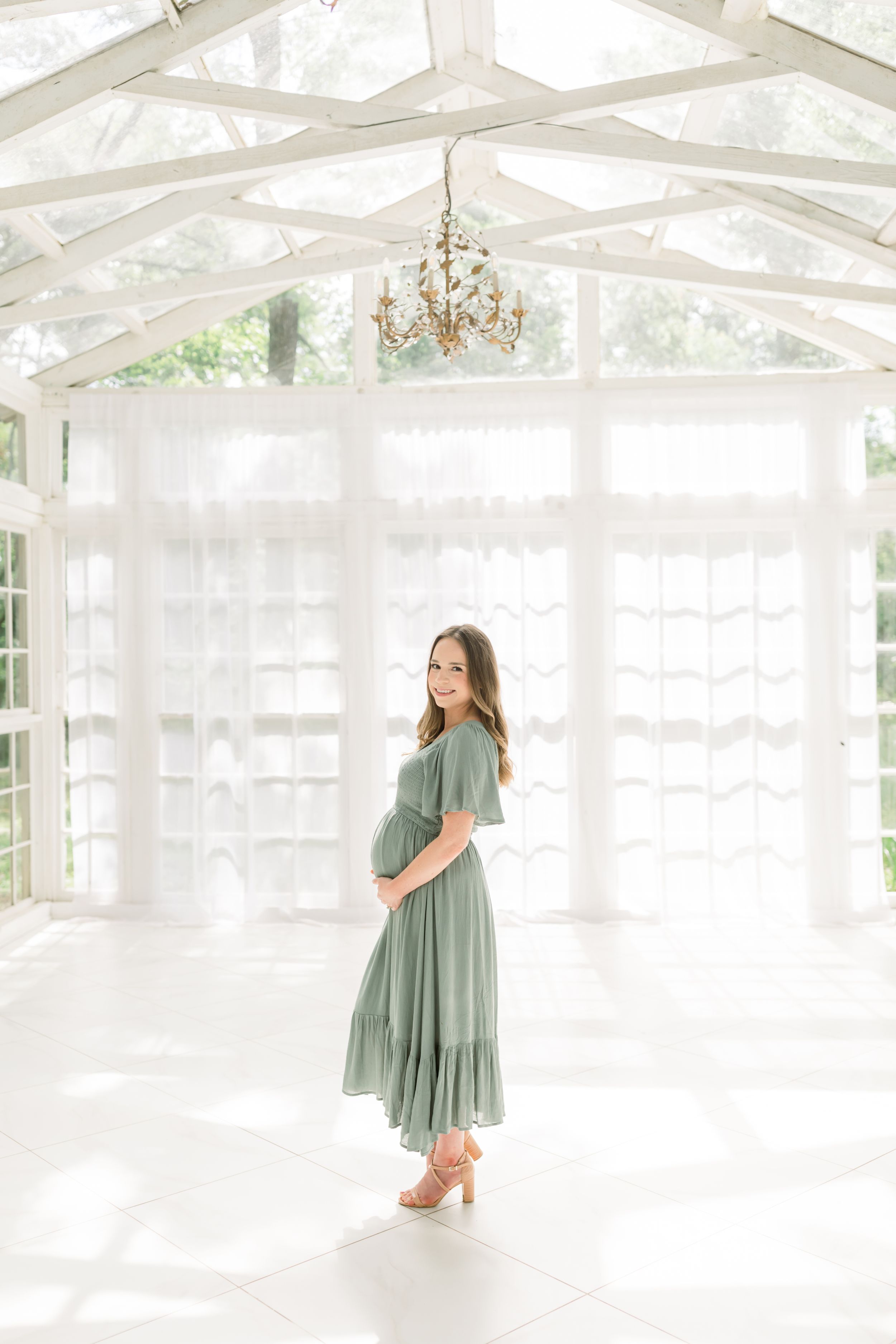 Expectant mother in sage green dress poses in an airy white sunroom with crystal chandelier.