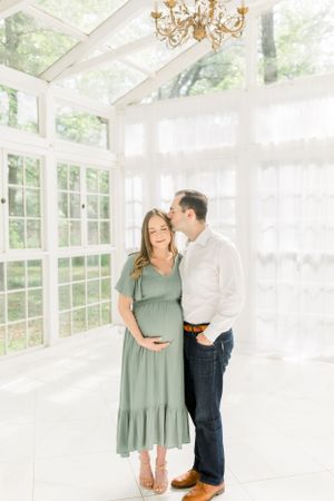 A tender maternity moment captured in a bright, airy sunroom with floor-to-ceiling windows.