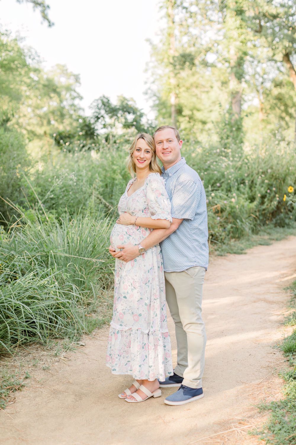 Couple poses outdoors on dirt path with tall grass, wearing coordinated light blue and white outfits.