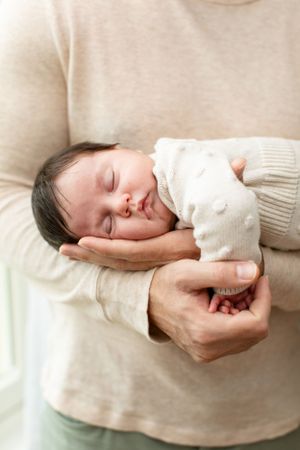 A sleeping newborn baby rests peacefully in gentle hands wearing a white knit sweater.