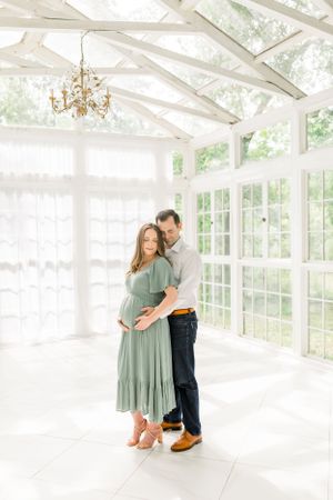 A tender maternity moment captured in a bright white conservatory with floor-to-ceiling windows.