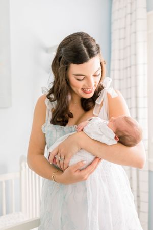 A woman in a light blue dress cradles a newborn baby in a bright nursery setting.