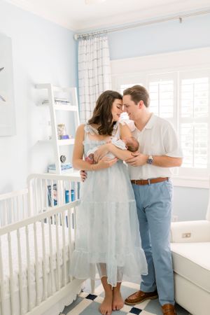 A couple stands together in a nursery holding their newborn baby near a white crib.