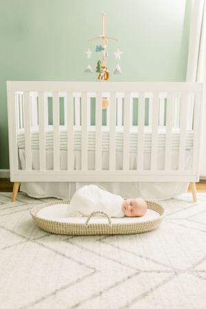 Modern white crib with mobile above sleeping newborn in bassinet on geometric rug.