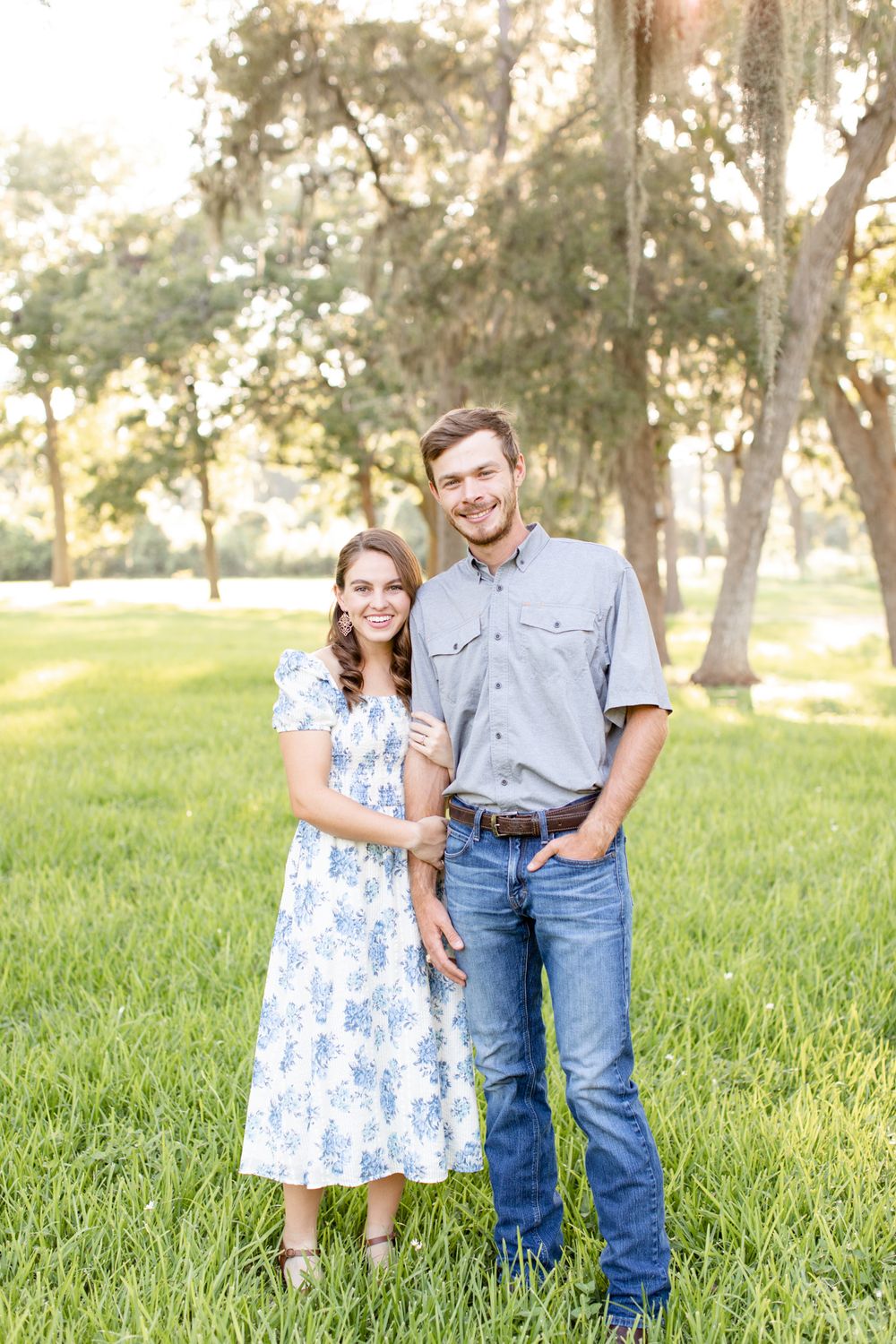 A couple in coordinating outfits poses together on a grassy lawn with Spanish moss trees behind them.