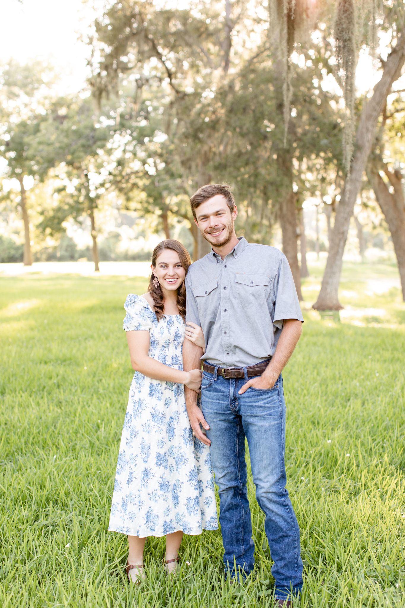 A couple in coordinating outfits poses together on a grassy lawn with Spanish moss trees behind them.