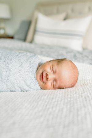 A sleeping newborn baby smiles while swaddled in gray on white bedding.