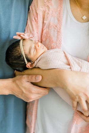 Close-up of sleeping newborn with pink bow headband being cradled in parent's arms.