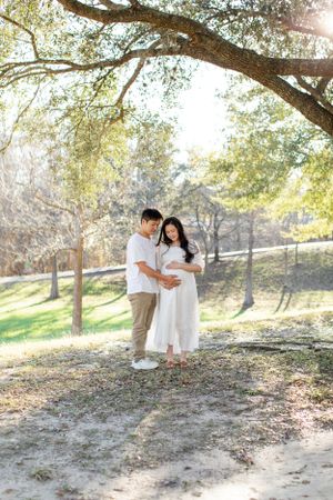 Maternity photo under sprawling tree branches in sunlit park.