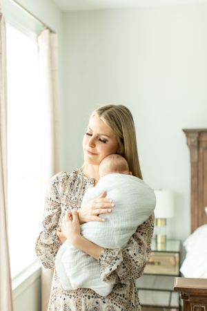 A woman in a floral dress cuddles her swaddled baby near a bright window.