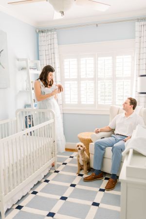 A nursery scene with parents, baby, and golden dog beside a white crib on checkered flooring.
