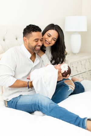 Couple sitting on bed sharing joyful moment with newborn baby wrapped in white blanket.