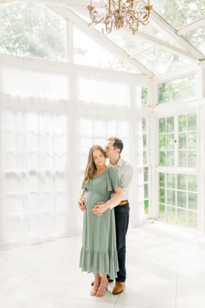 A loving maternity portrait in a sunlit conservatory with white curtains and chandelier.