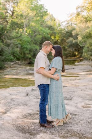 A couple embraces in a romantic moment on rocky terrain surrounded by lush green trees in golden sunlight.