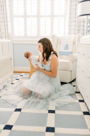 A woman in a flowing dress sits on a checkered floor holding a baby in a bright nursery.