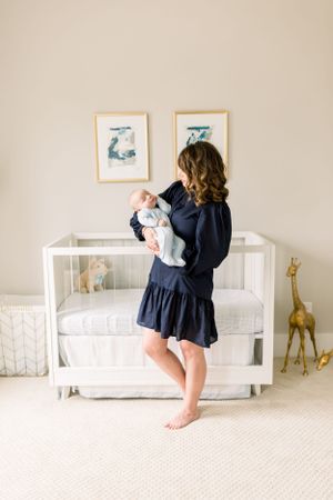 Parent in navy dress holds infant beside white crib with giraffe decor and framed art.