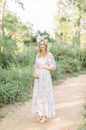 Expectant mother in floral maxi dress standing on nature path surrounded by greenery.