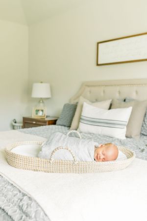 A sleeping newborn baby lies in a woven basket on a gray and white bed.