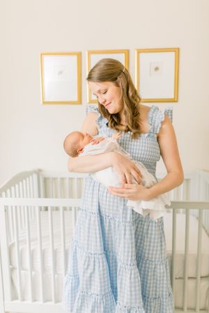 A blue gingham dress-clad figure cradles a newborn in a peaceful nursery setting.