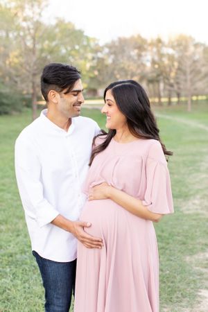 Expecting couple in white and pink outfits sharing tender moment outdoors.