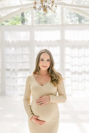 Intimate maternity portrait in a sun-drenched white room with crystal chandelier overhead.
