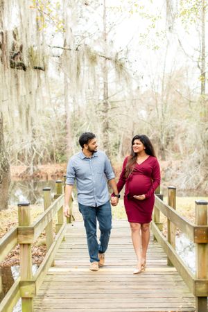 Couple walks hand in hand on wooden boardwalk through misty forest during maternity photo session.
