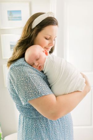 Tender embrace of newborn wrapped in white against blue floral dress.