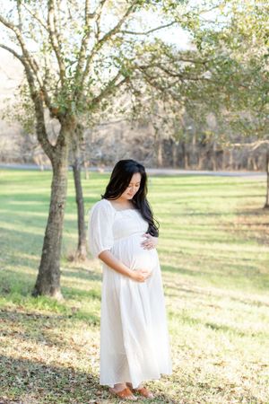 Expecting mother in white dress stands alone in serene park setting.