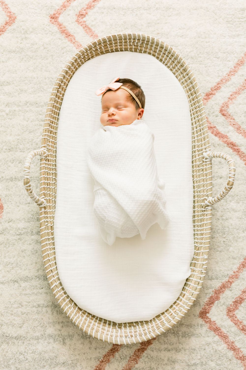 Sleeping newborn swaddled in white blanket rests in woven bassinet on patterned rug.
