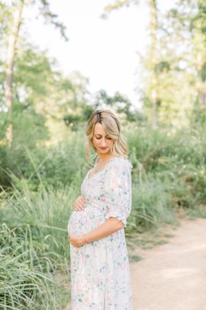 Expectant mother in floral maxi dress poses on nature trail surrounded by lush greenery.