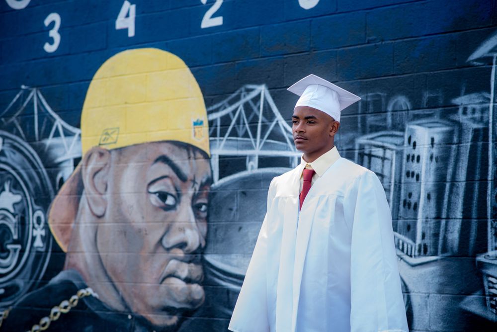 Graduate in white cap and gown standing in front of urban mural art.