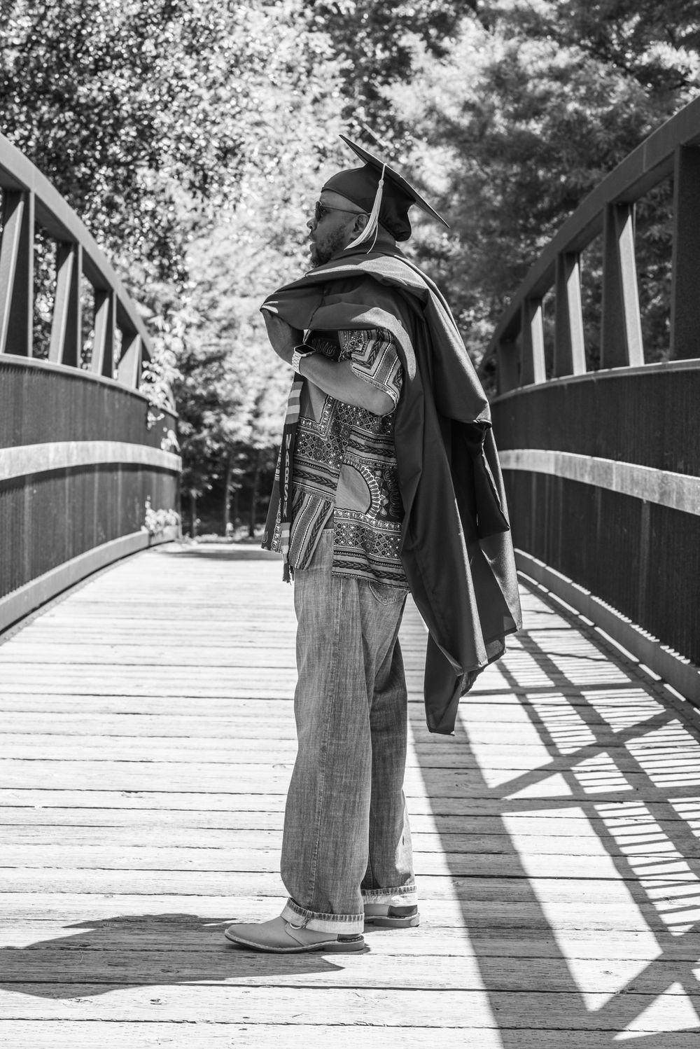 Black and white photo of person in graduation attire standing on wooden bridge.