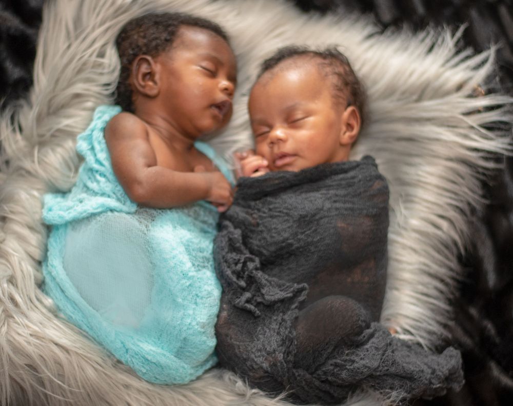 Twin newborns in turquoise and grey outfits sleeping together on a furry blanket.