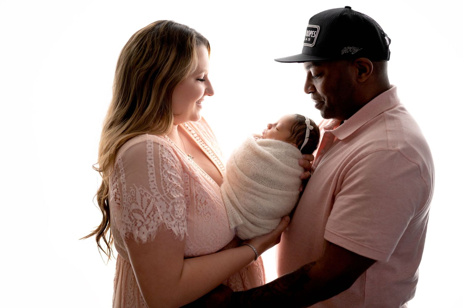 Tender moment between couple holding newborn baby wrapped in white against light background.