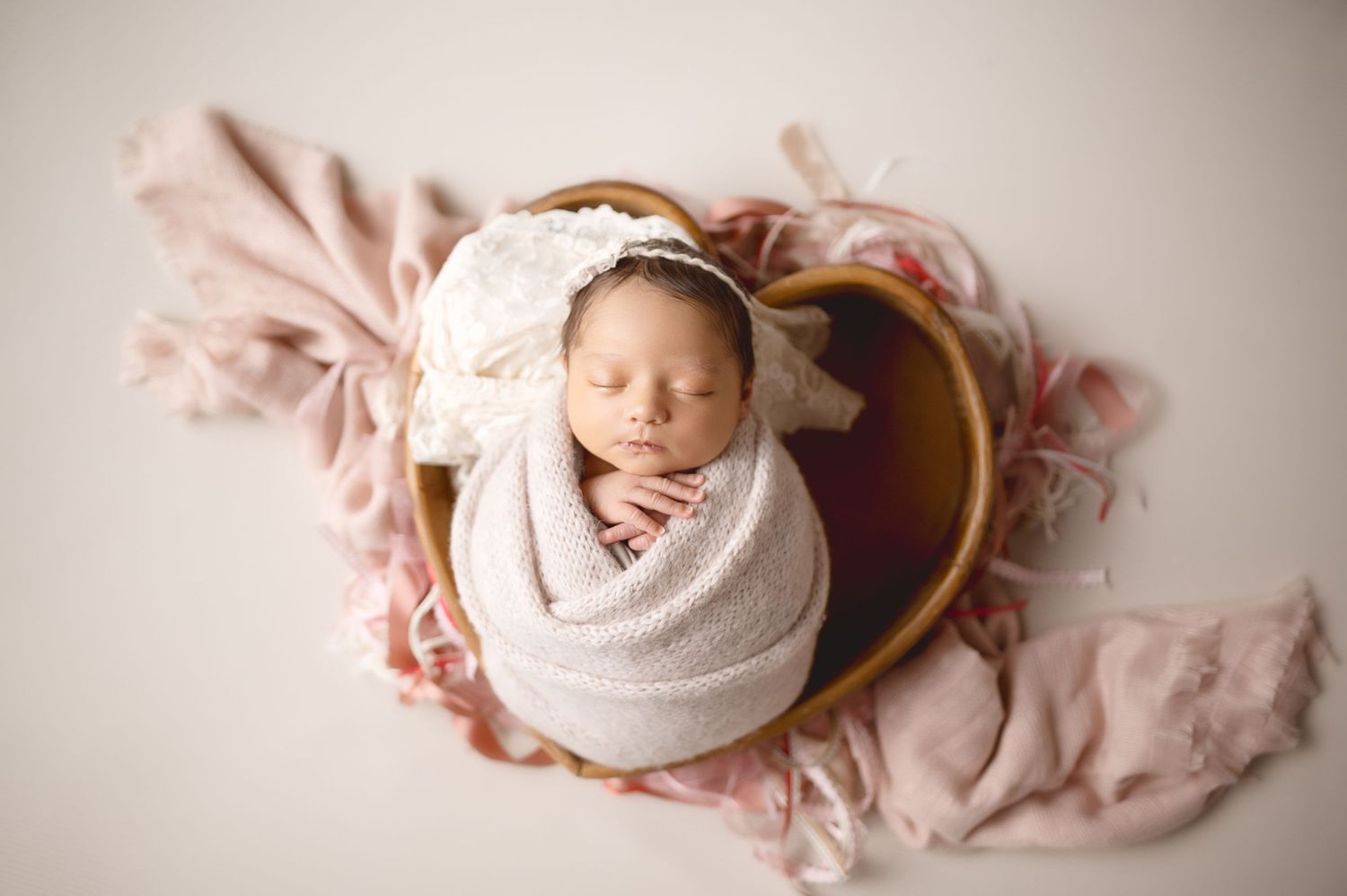Sleeping newborn in white wrap nestled in heart-shaped basket with pink fabric accents.