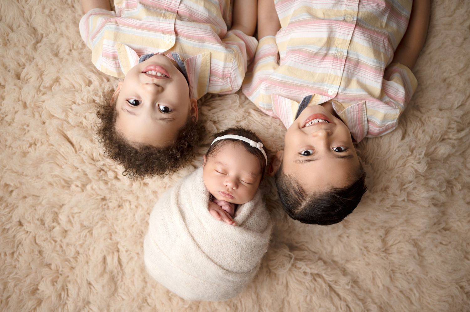 Three siblings lying on cream carpet with newborn baby in center wearing matching outfits.